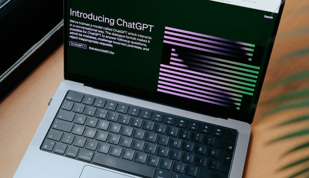 A sleek laptop on a wooden desk showing the ChatGPT homepage by a window.