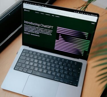 A sleek laptop on a wooden desk showing the ChatGPT homepage by a window.
