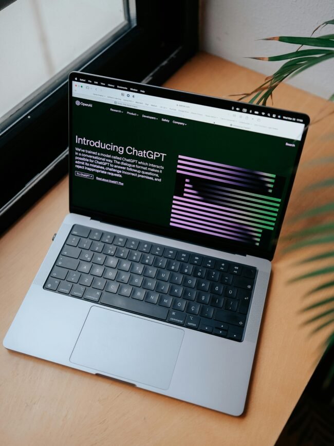 A sleek laptop on a wooden desk showing the ChatGPT homepage by a window.